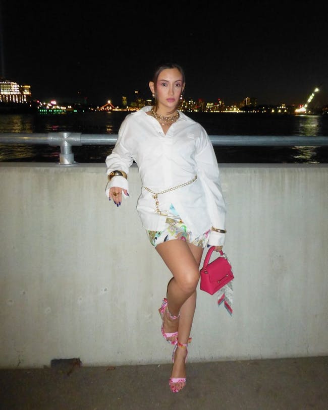 A woman in a white dress and pink purse leaning against a wall