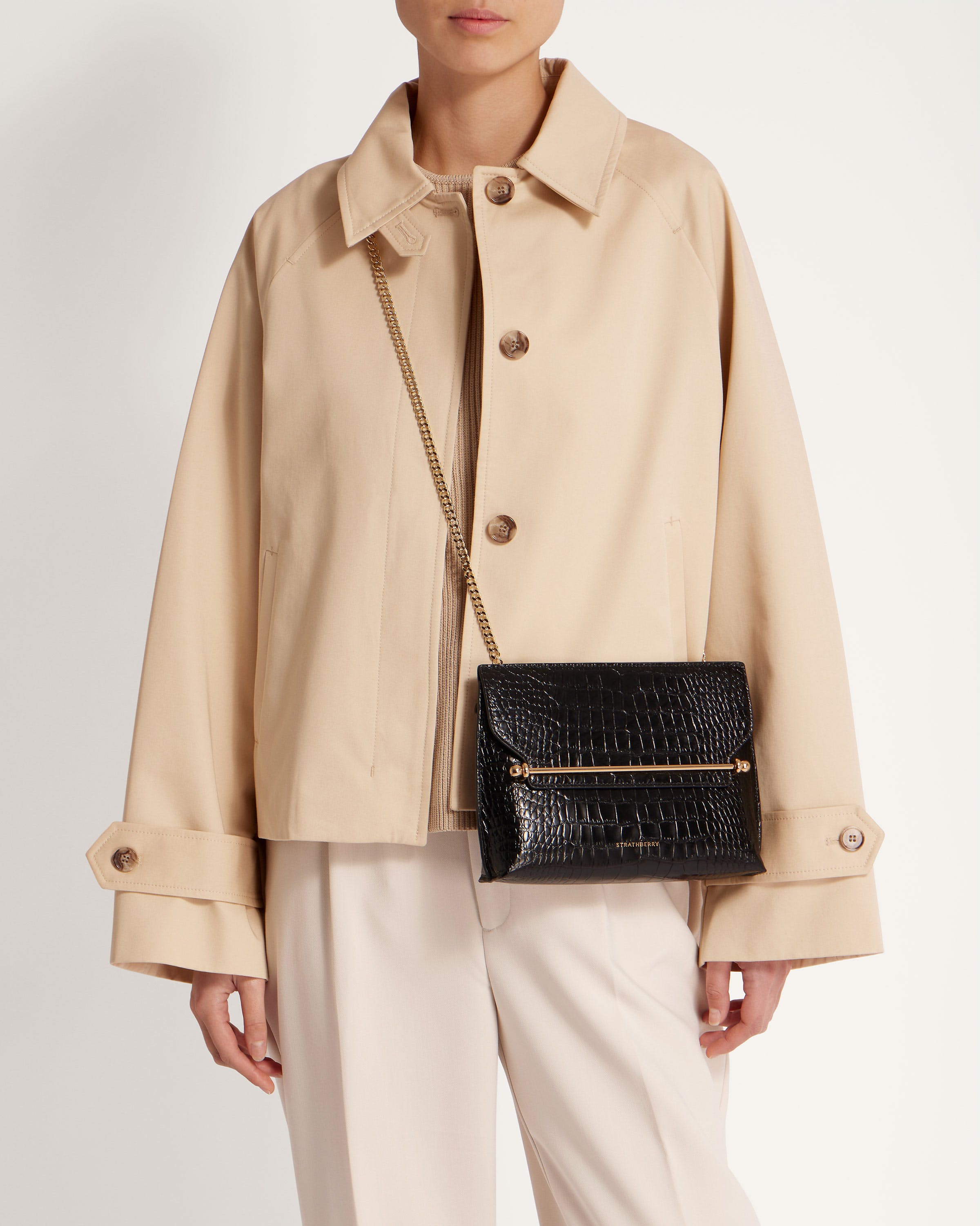 A woman in a beige coat and white pants holding a black purse