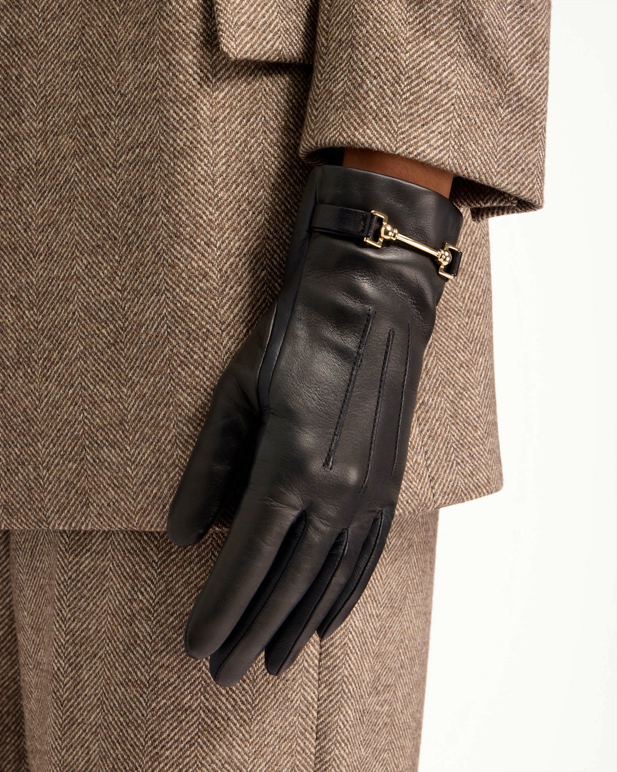 Black leather glove with gold horsebit detail, worn with brown herringbone coat.