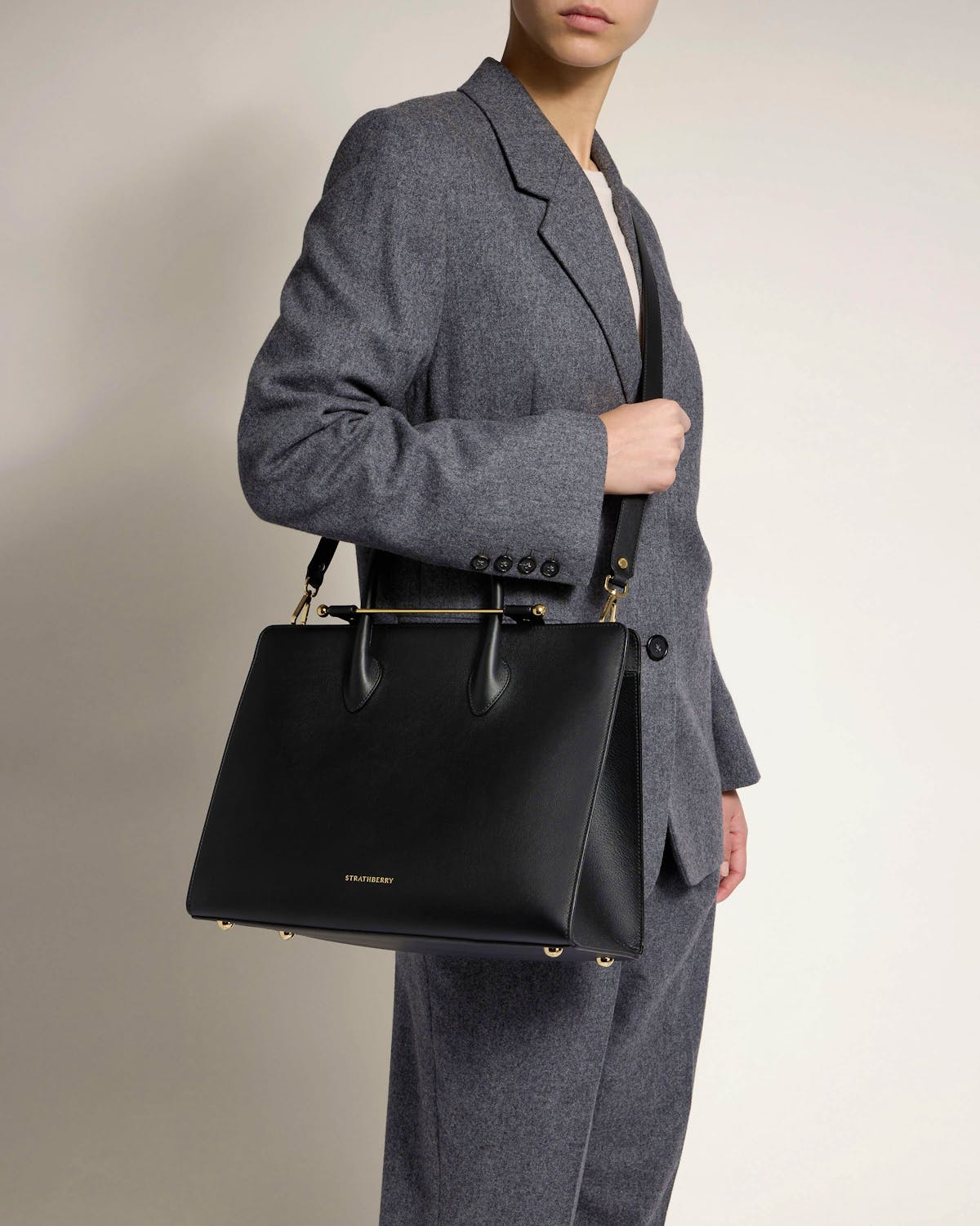 Model in grey suit holding black Strathberry tote crossbody at hip