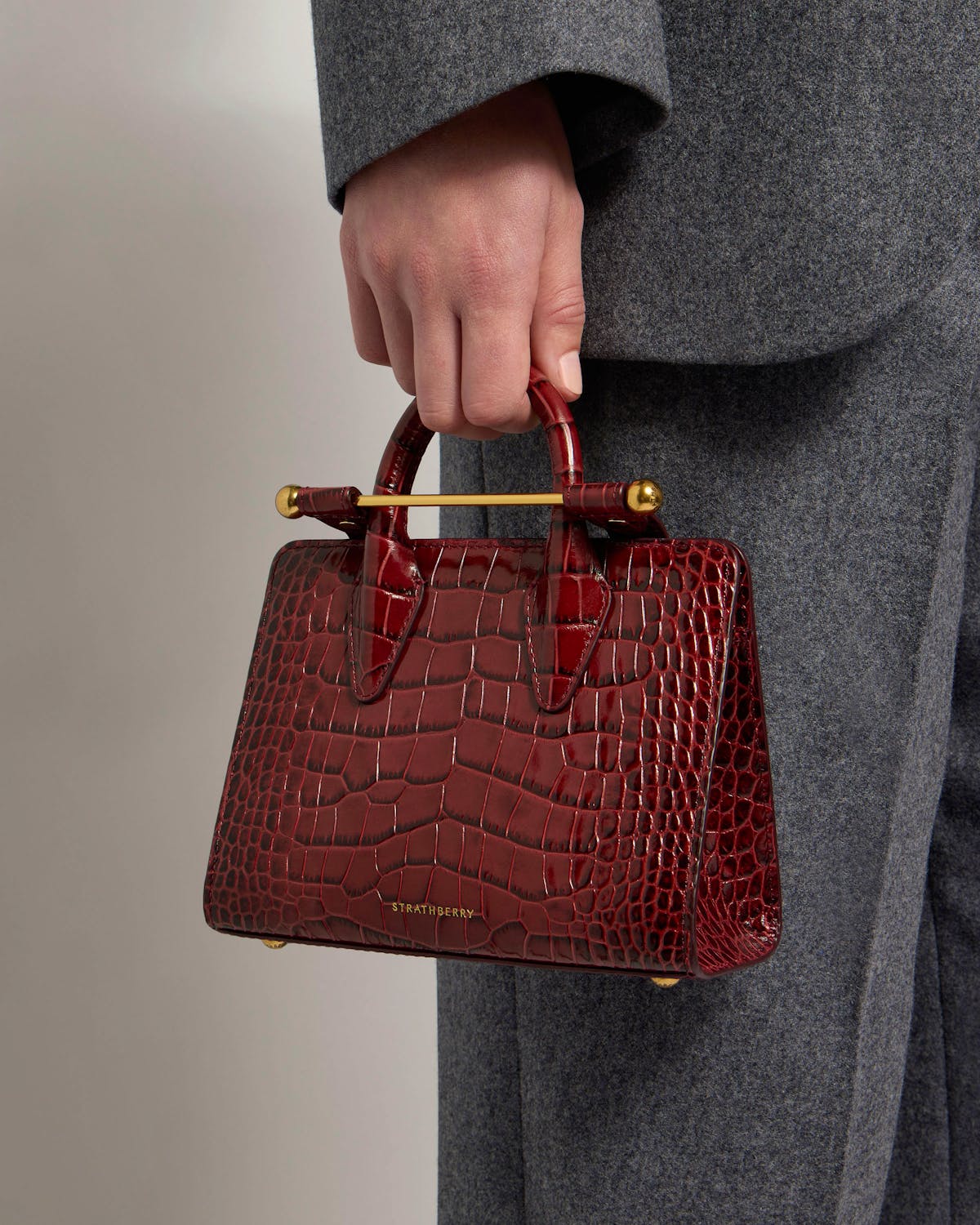 Strathberry croc nano tote in burgundy held by top handle at hip.