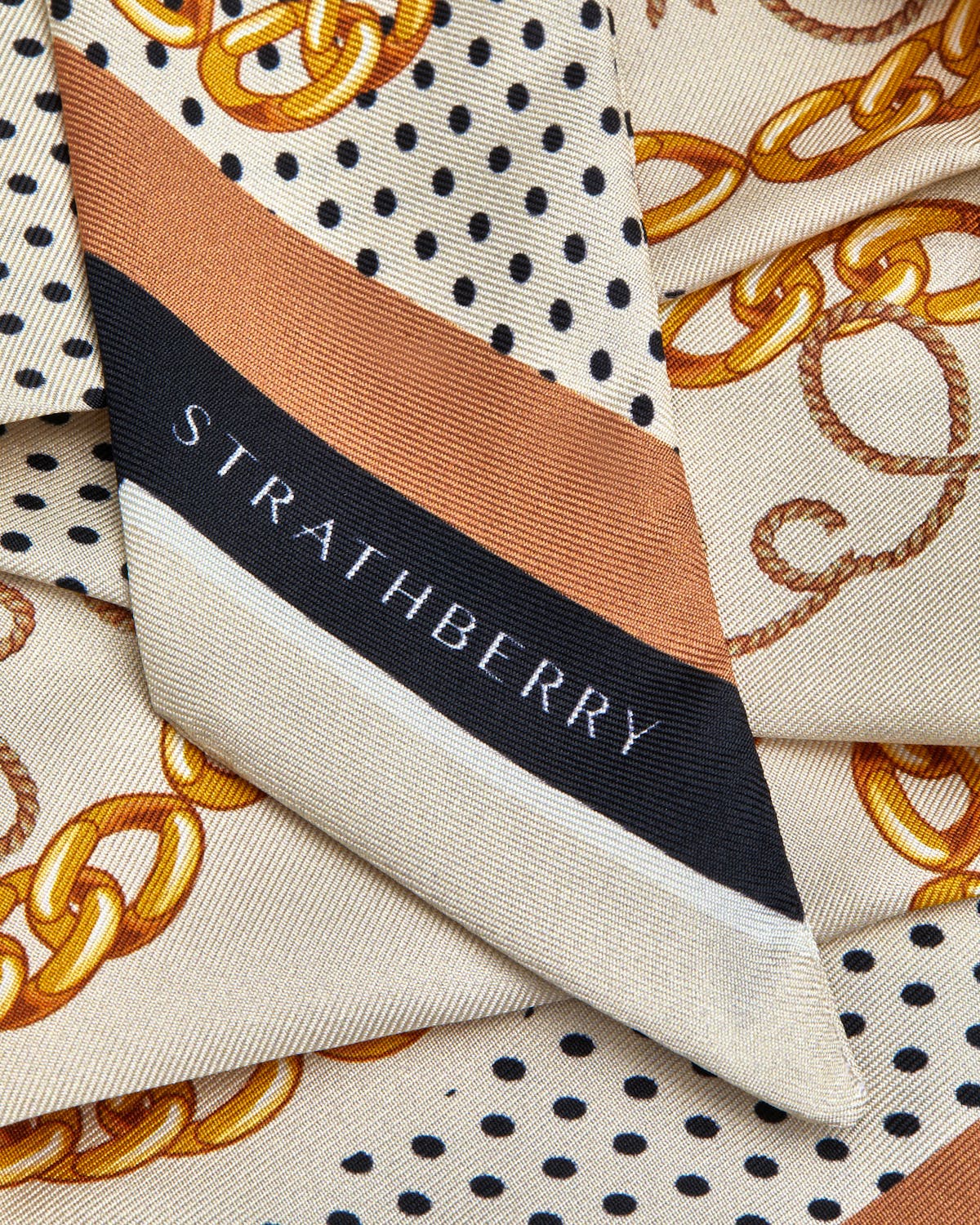Folded Strathberry silk skinny scarf showing tan chain print and black polka dots.