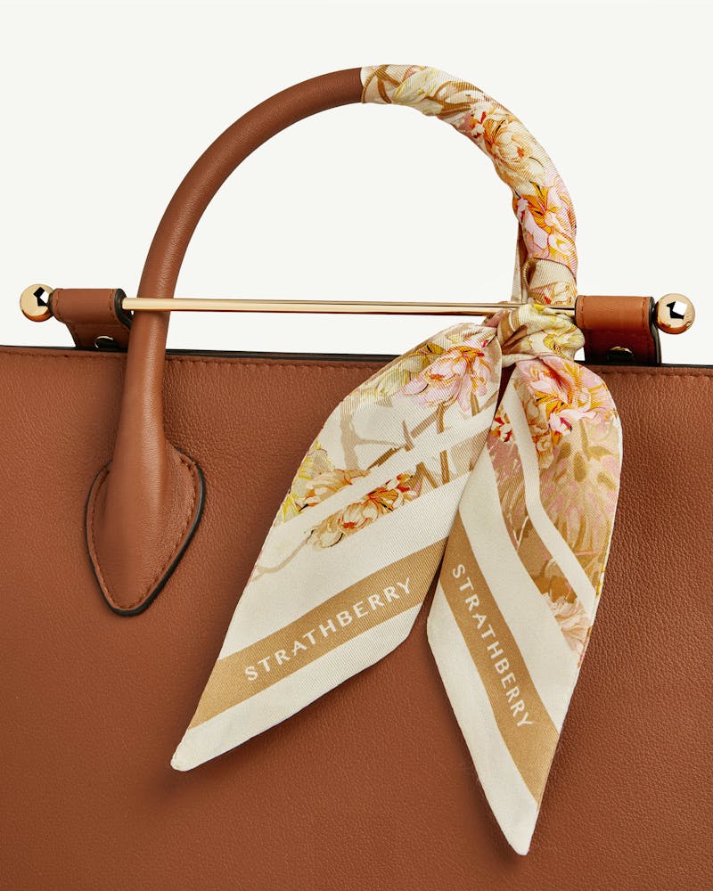 Vanilla and pink floral silk skinny scarf tied to brown leather handbag handle, Strathberry.
