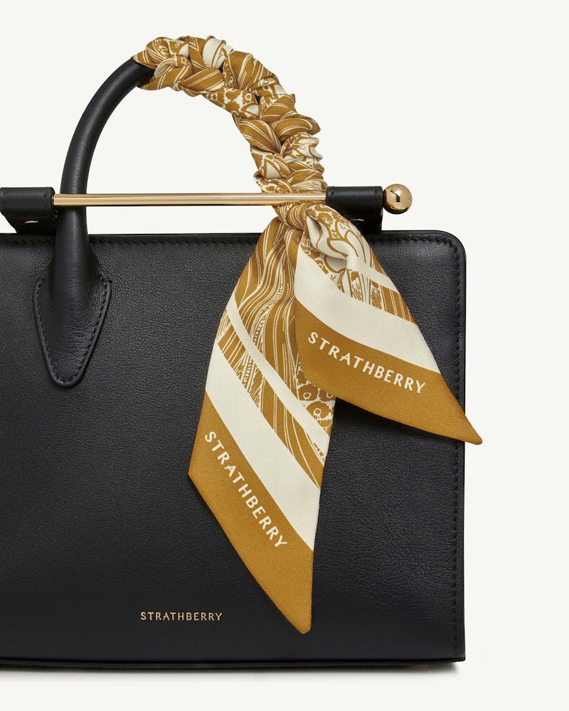 Black leather bag with mustard wallpaper-print silk skinny scarf tied to handle, Strathberry logo