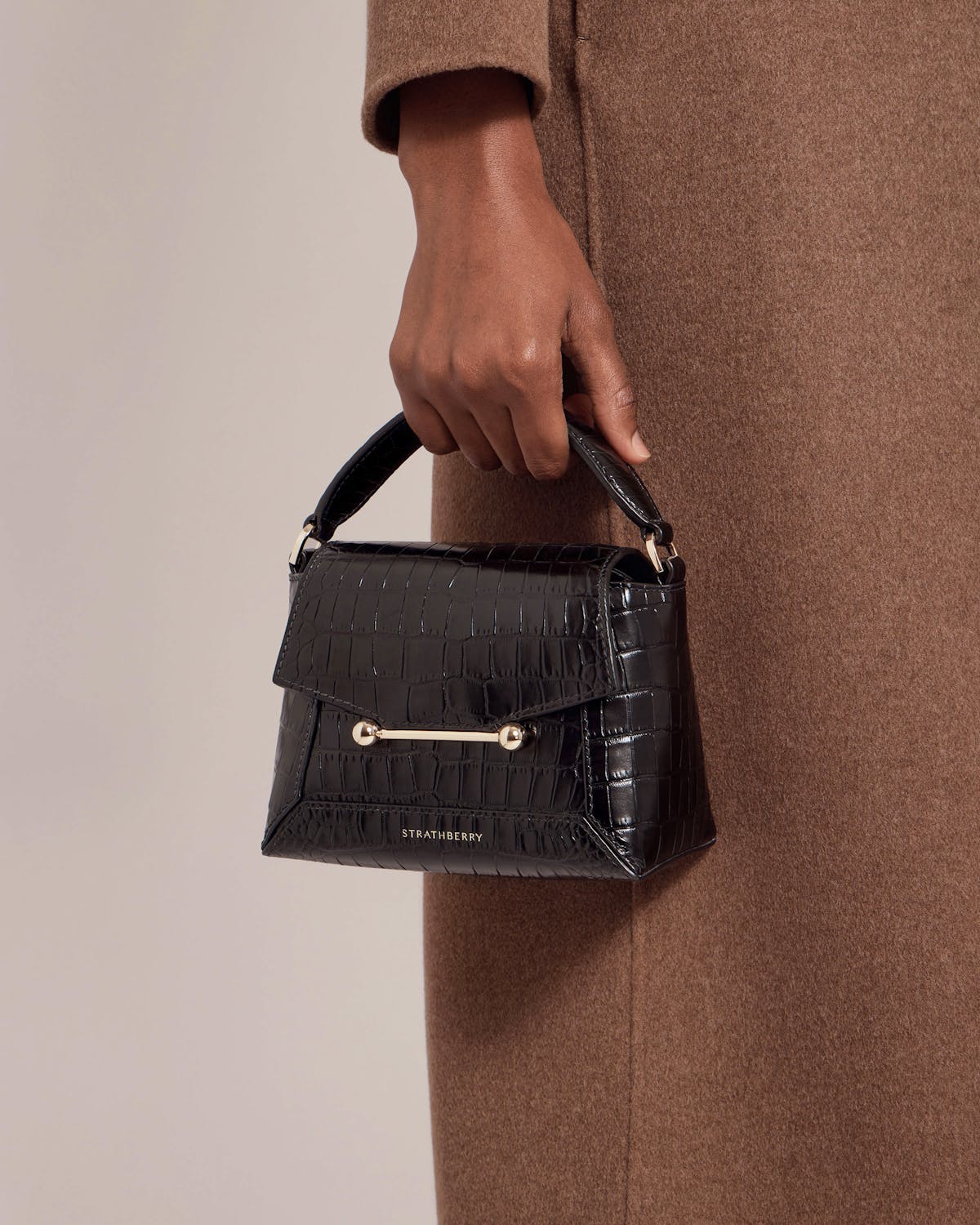 Strathberry Mosaic Nano soft croc top-handle bag held at hip against brown coat