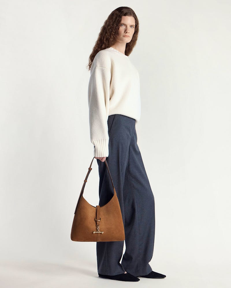 Model in white sweater and grey trousers holding tan suede kite hobo bag.