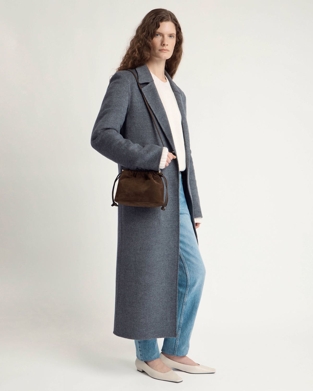 Model wearing long grey coat and jeans carrying chocolate suede drawstring bag.