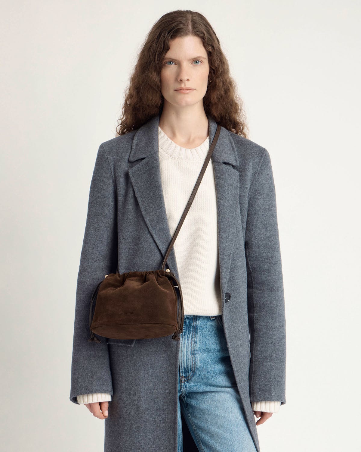 Model wearing a grey coat and jeans with a chocolate suede crossbody drawstring bag.