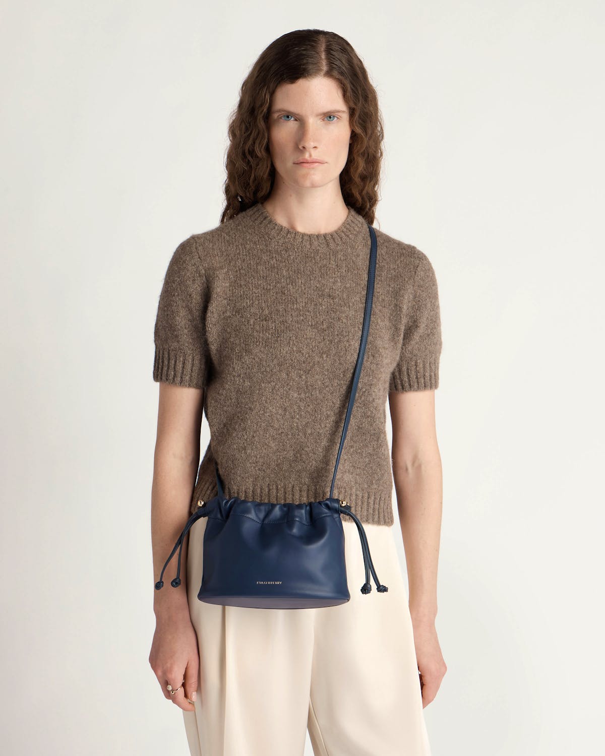 Model wearing brown sweater and cream trousers with marine blue drawstring crossbody bag.