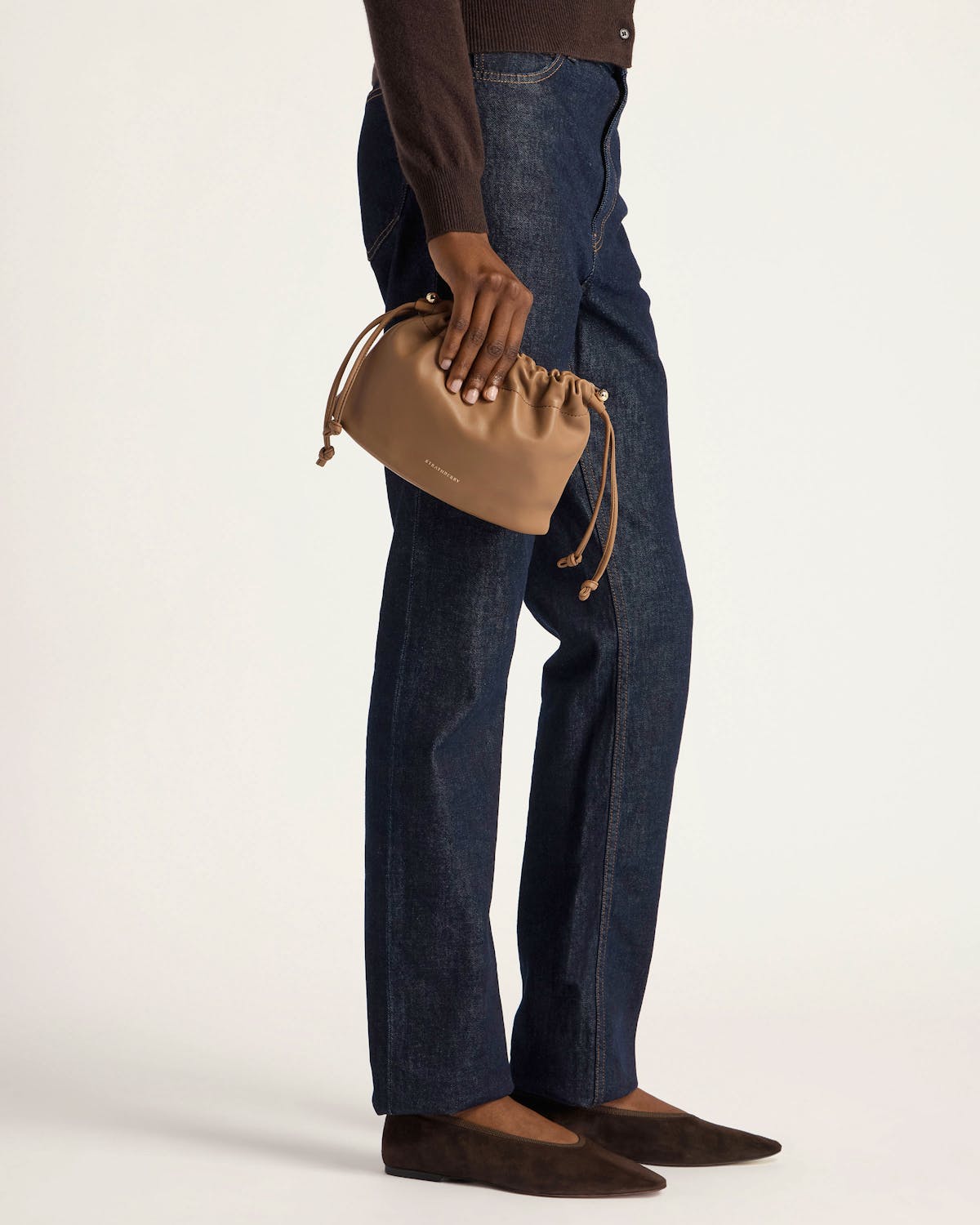Model holding clay Charlotte drawstring bag at side, wearing dark jeans and brown shoes