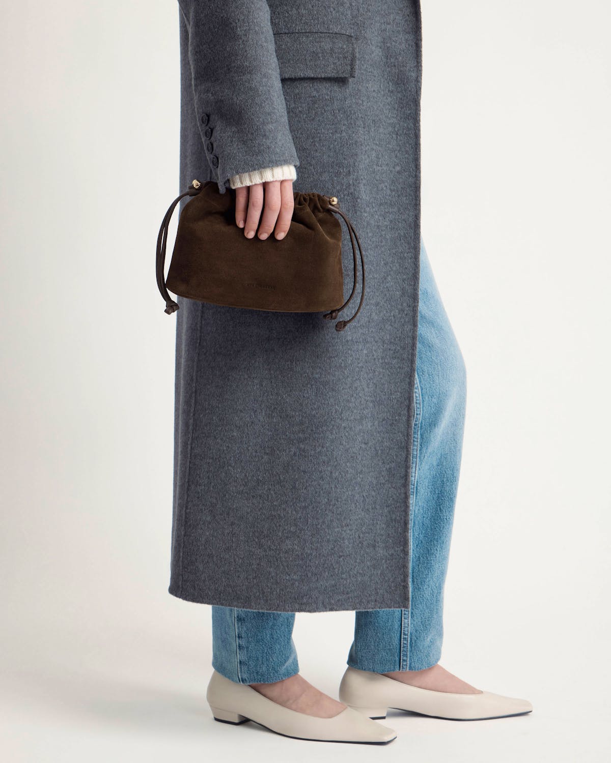 Model holding chocolate suede drawstring bag with grey coat, jeans, cream flats.