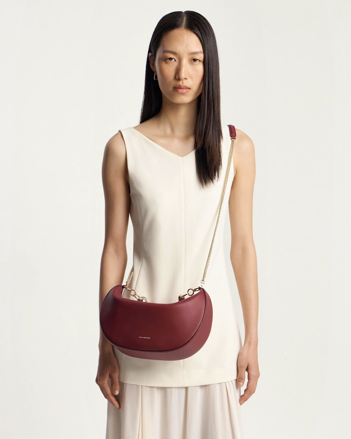 Model wearing white sleeveless dress carrying oxblood crescent moon bag with gold chain strap.