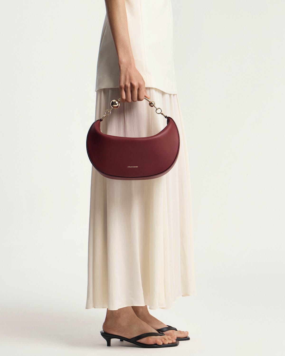 Model holding oxblood crescent moon bag with gold chain handle, Strathberry logo.