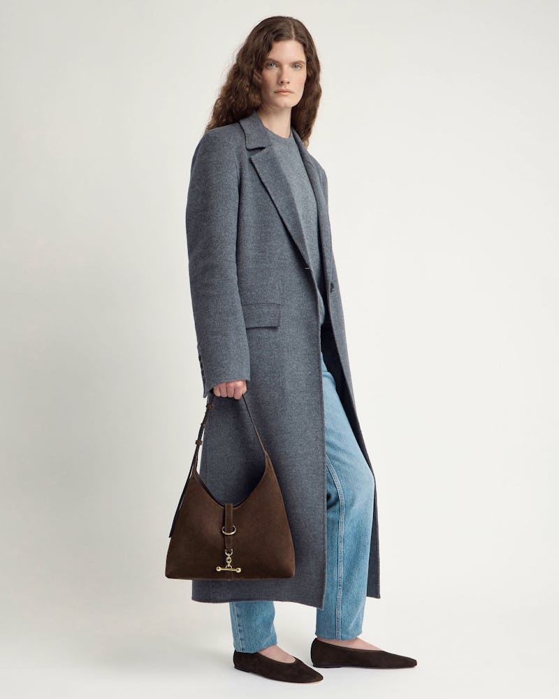 Model wearing grey coat holding chocolate suede Kite Hobo bag with gold hardware.