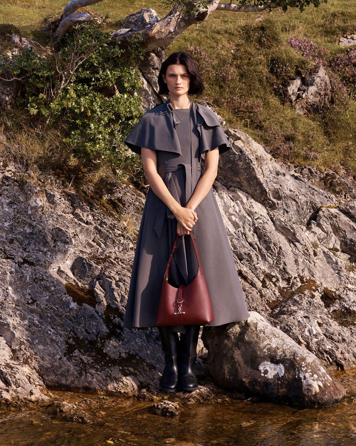 Model standing on rocks holding an oxblood Kite Hobo bag.