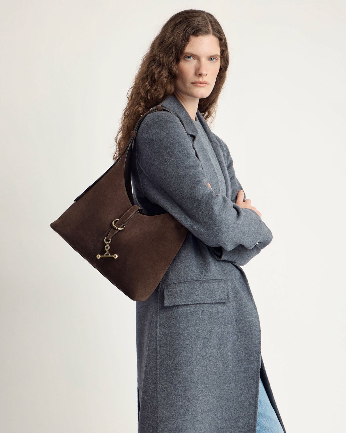 Model wearing grey coat carrying chocolate suede kite hobo bag with gold hardware.