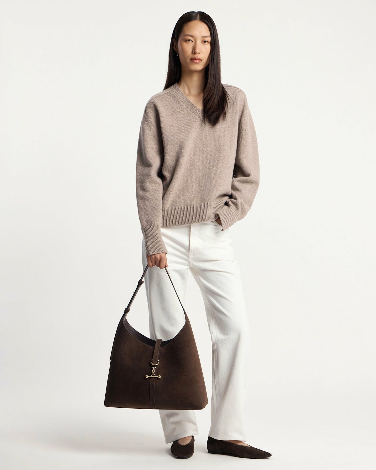 Model wearing beige V‑neck jumper and white trousers holding chocolate suede hobo bag.