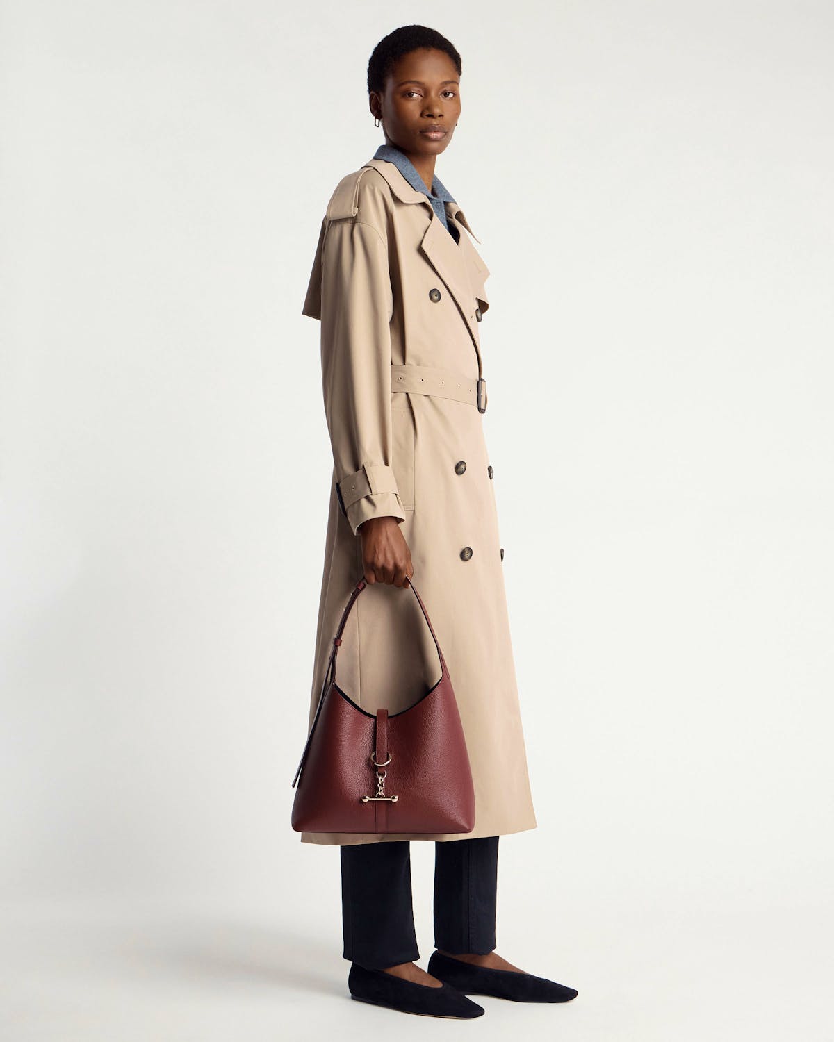 Model wearing beige trench coat holding an oxblood Kite Hobo bag with metal ring detail.