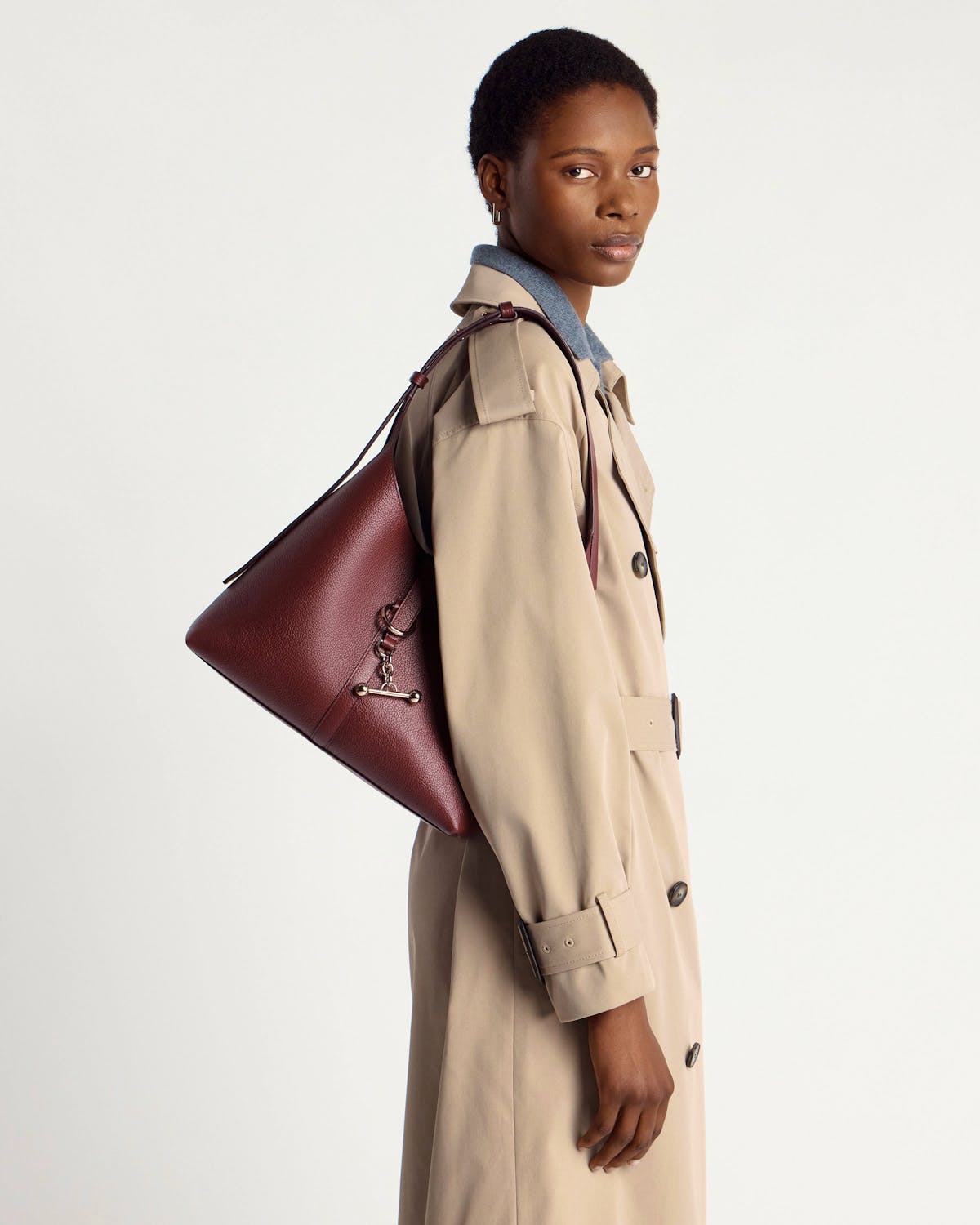 Model wearing an oxblood Kite Hobo bag on shoulder, trench coat side view.