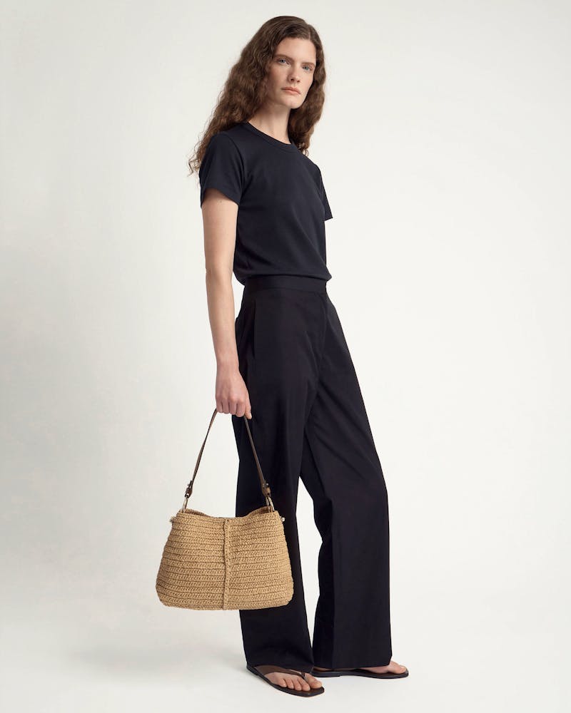 Woman wearing black outfit holding a woven raffia hobo bag with clay-coloured strap.