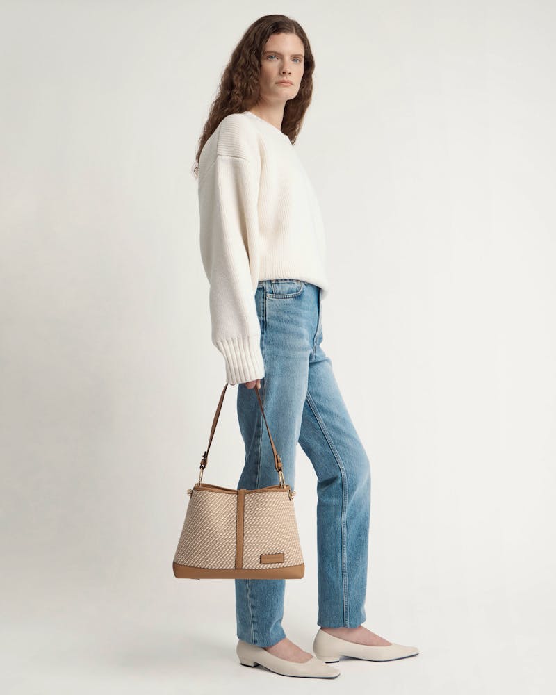Model standing in white jumper and jeans holding caramel and natural twill hobo bag.