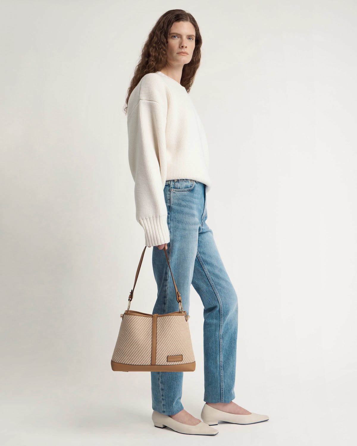 Model standing in white jumper and jeans holding caramel and natural twill hobo bag.