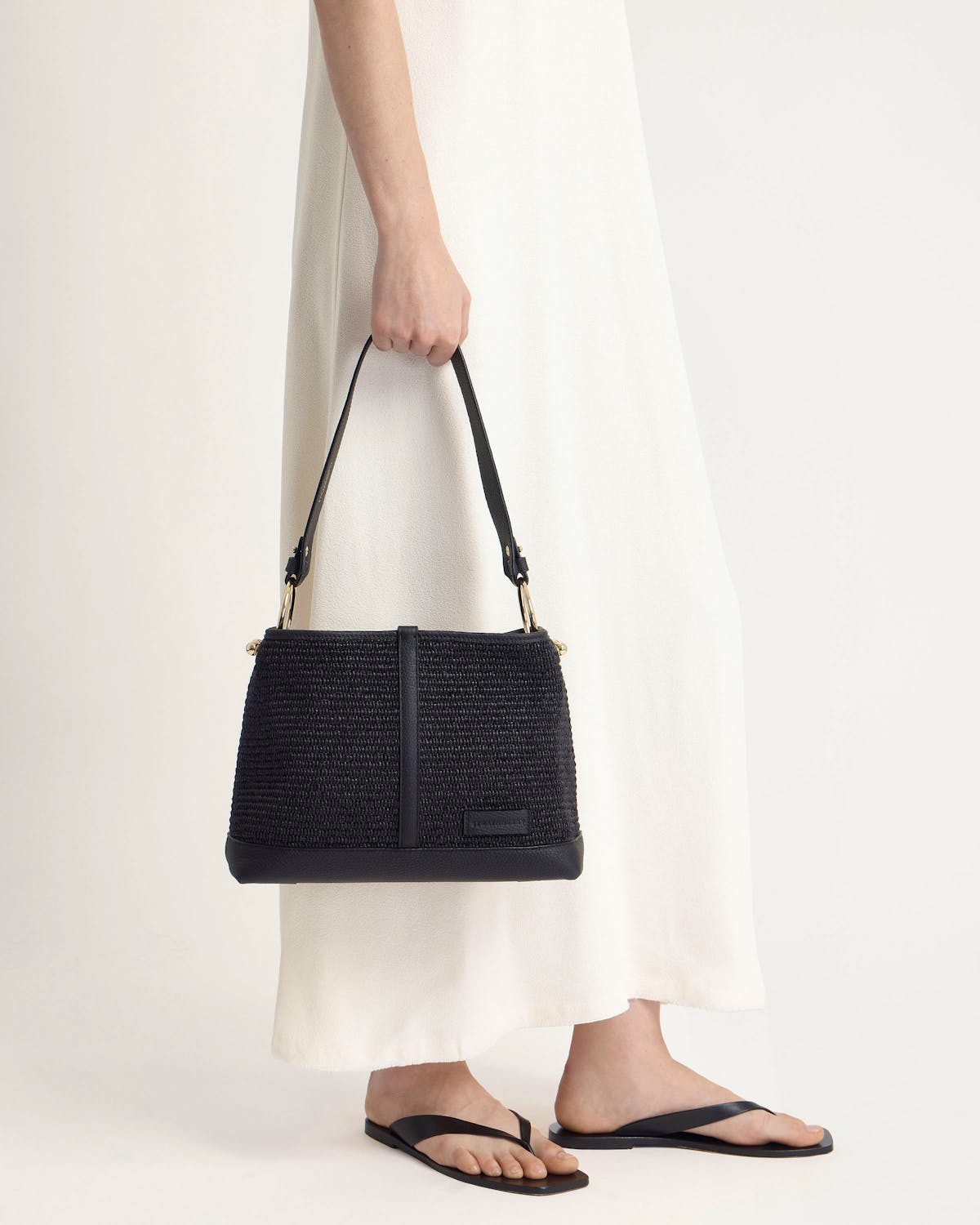 Woman holding a black raffia hobo bag against a white dress.