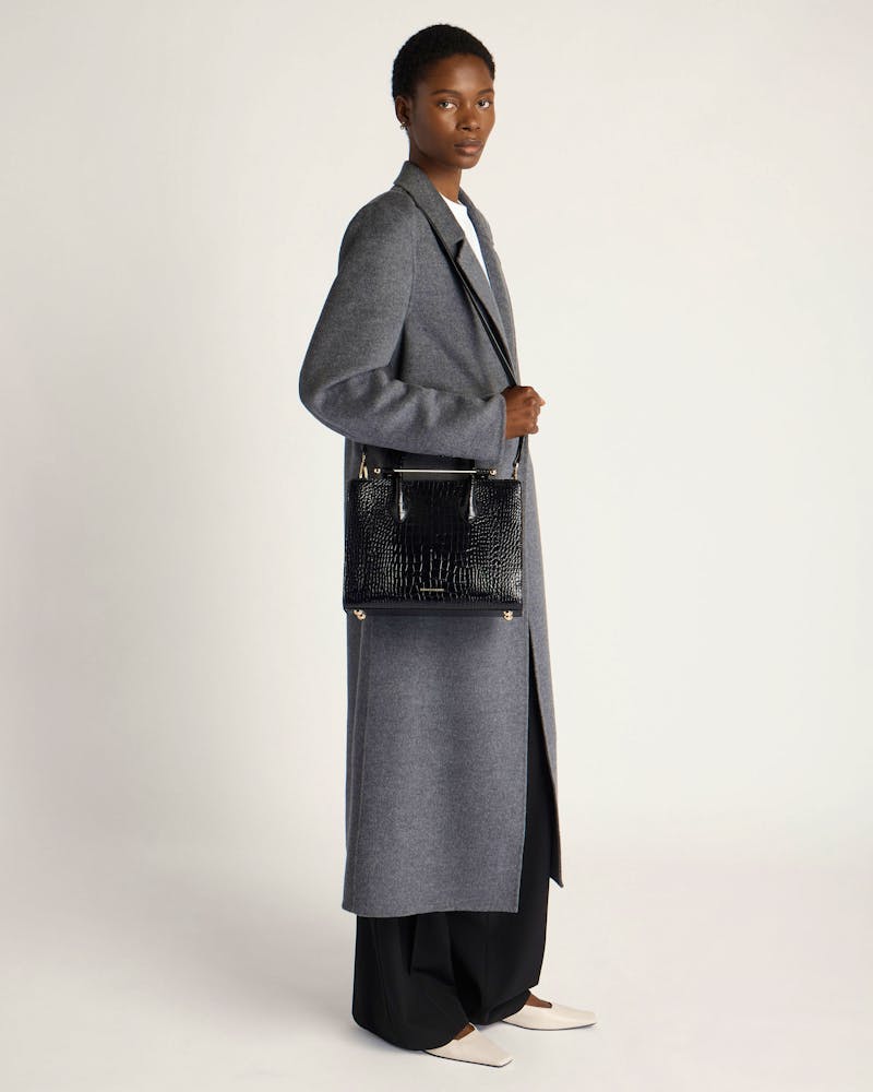 Model wearing grey long coat holding black croc midi tote bag at side.