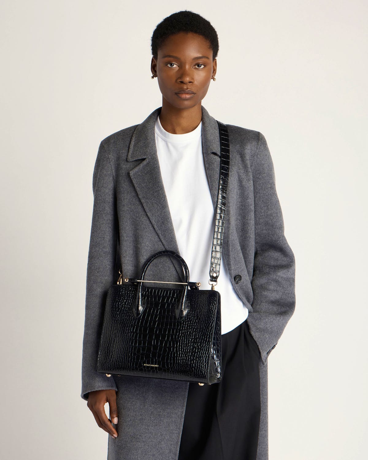 Model wearing grey coat carrying Strathberry black croc midi tote with shoulder strap.