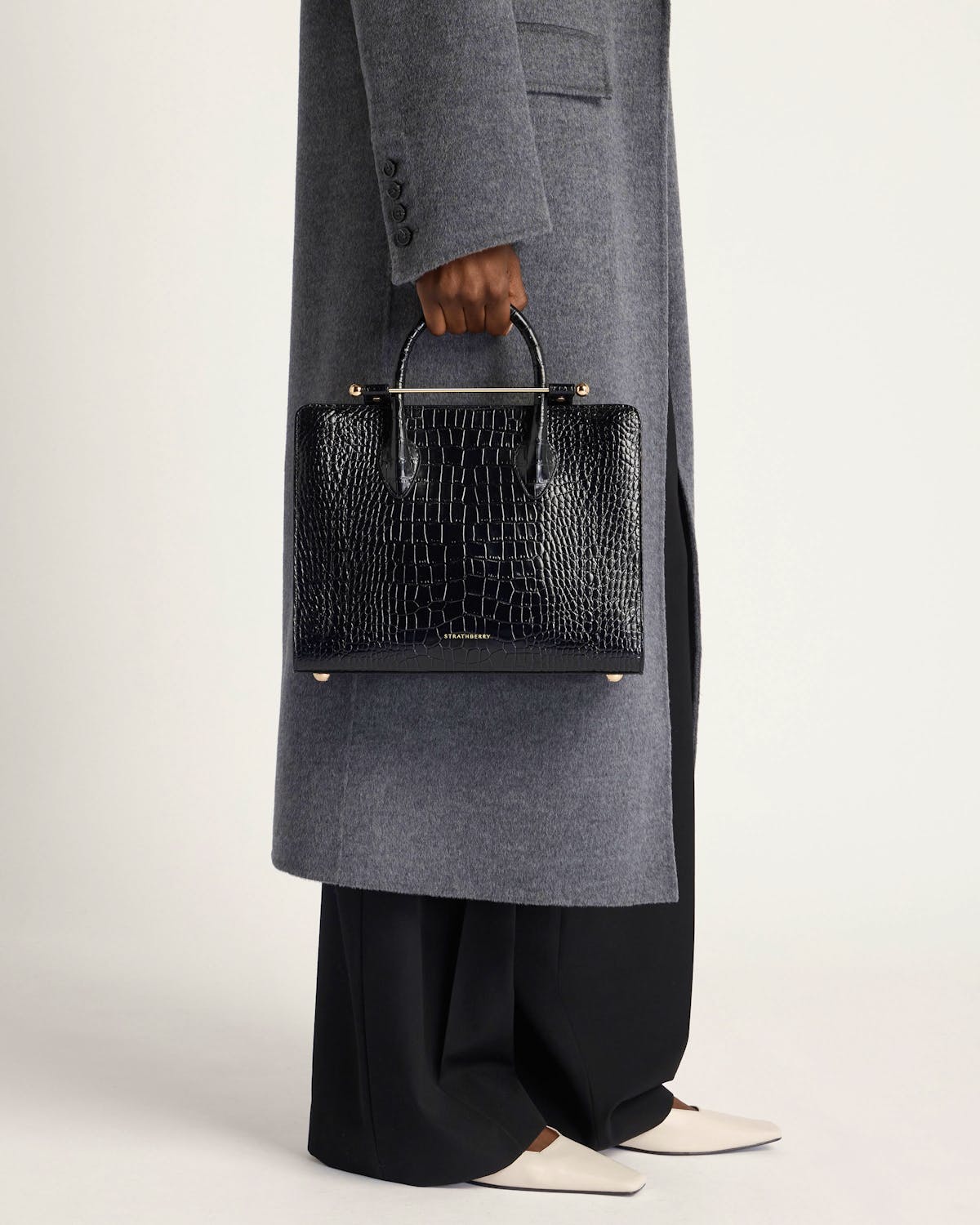 Model holding Strathberry midi tote in black crocodile-embossed leather, worn with grey coat.