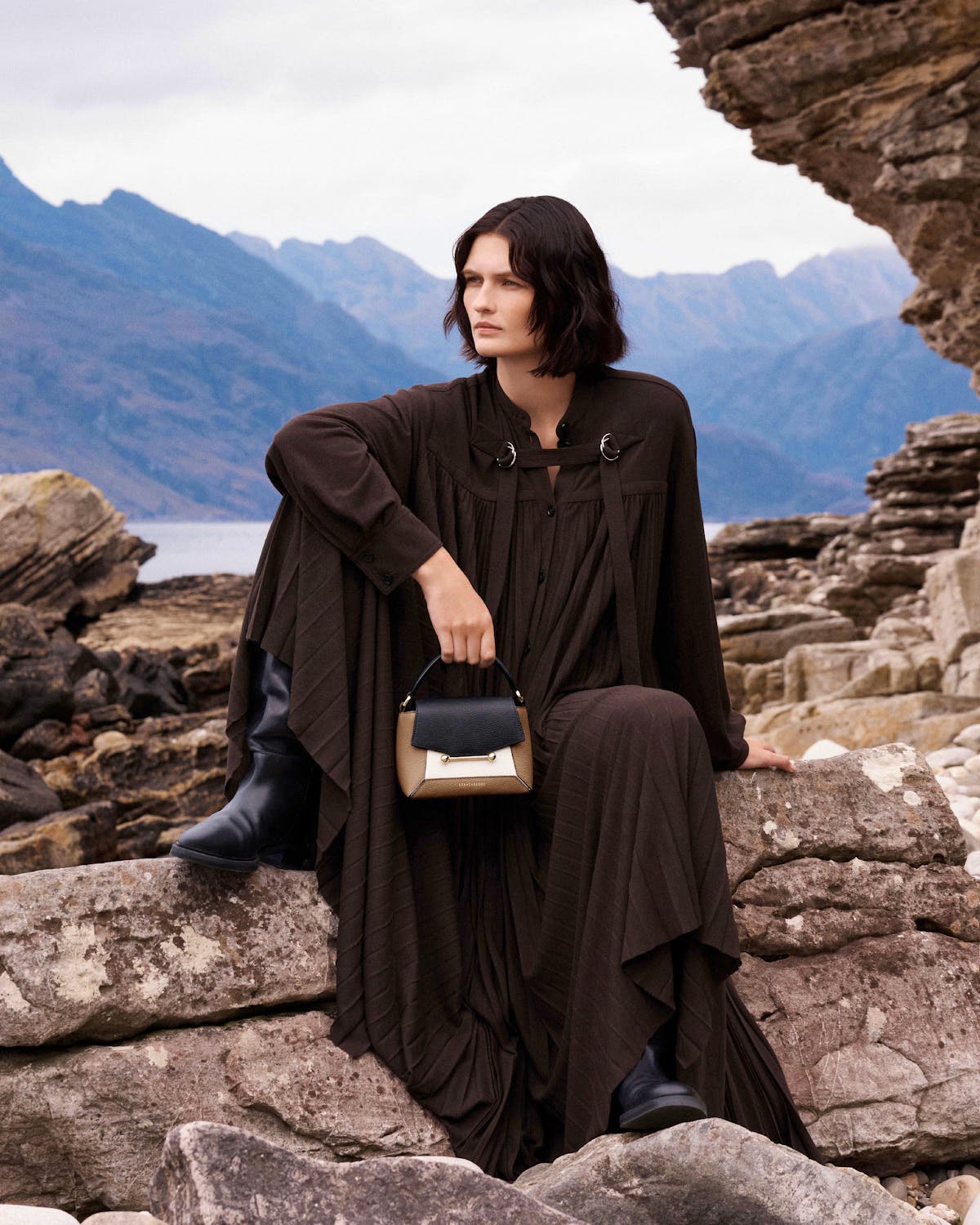 Model seated on rocks holding a clay, black and white small top-handle bag.