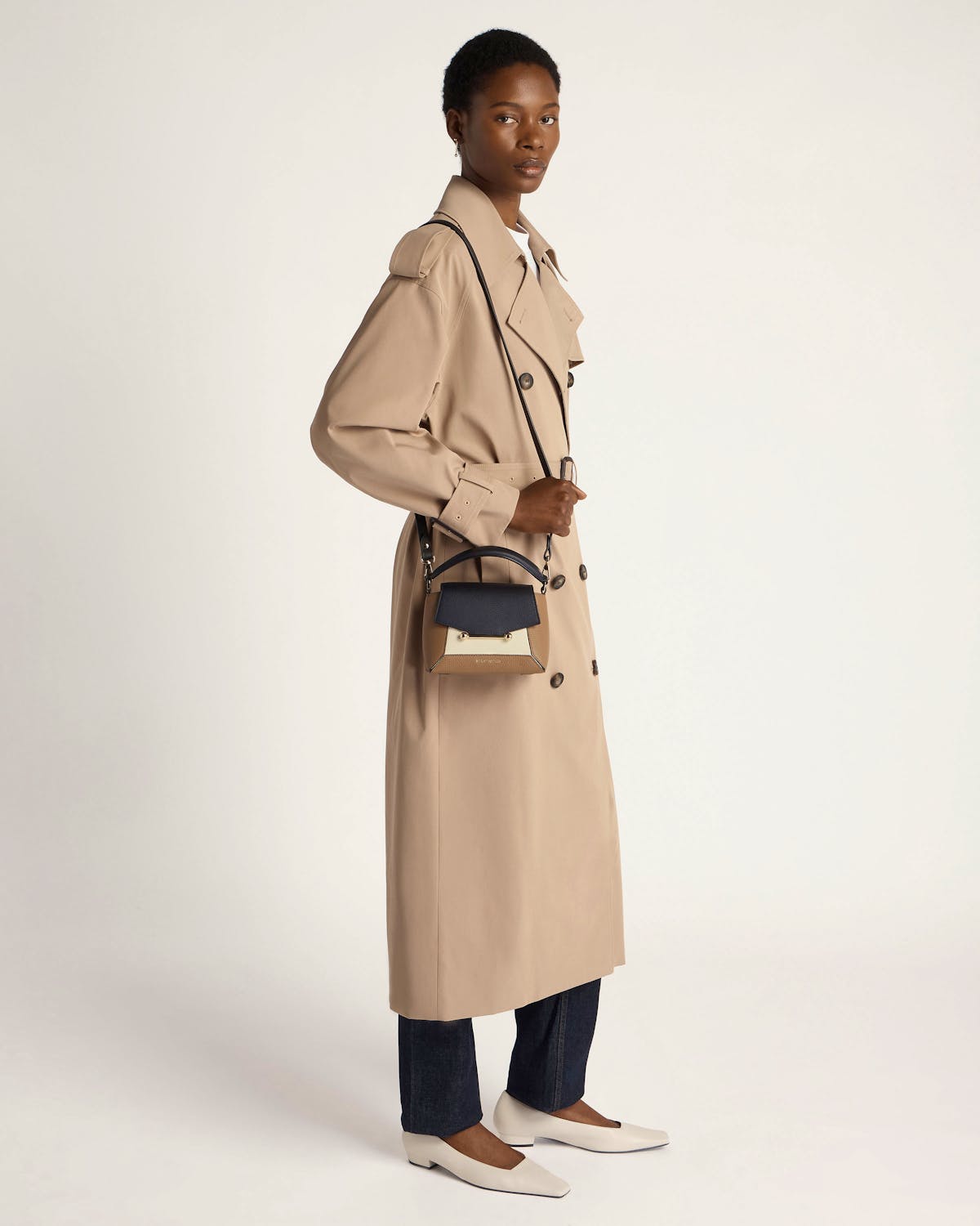 Model wearing beige trench coat carrying a clay, black and white cross-body bag.