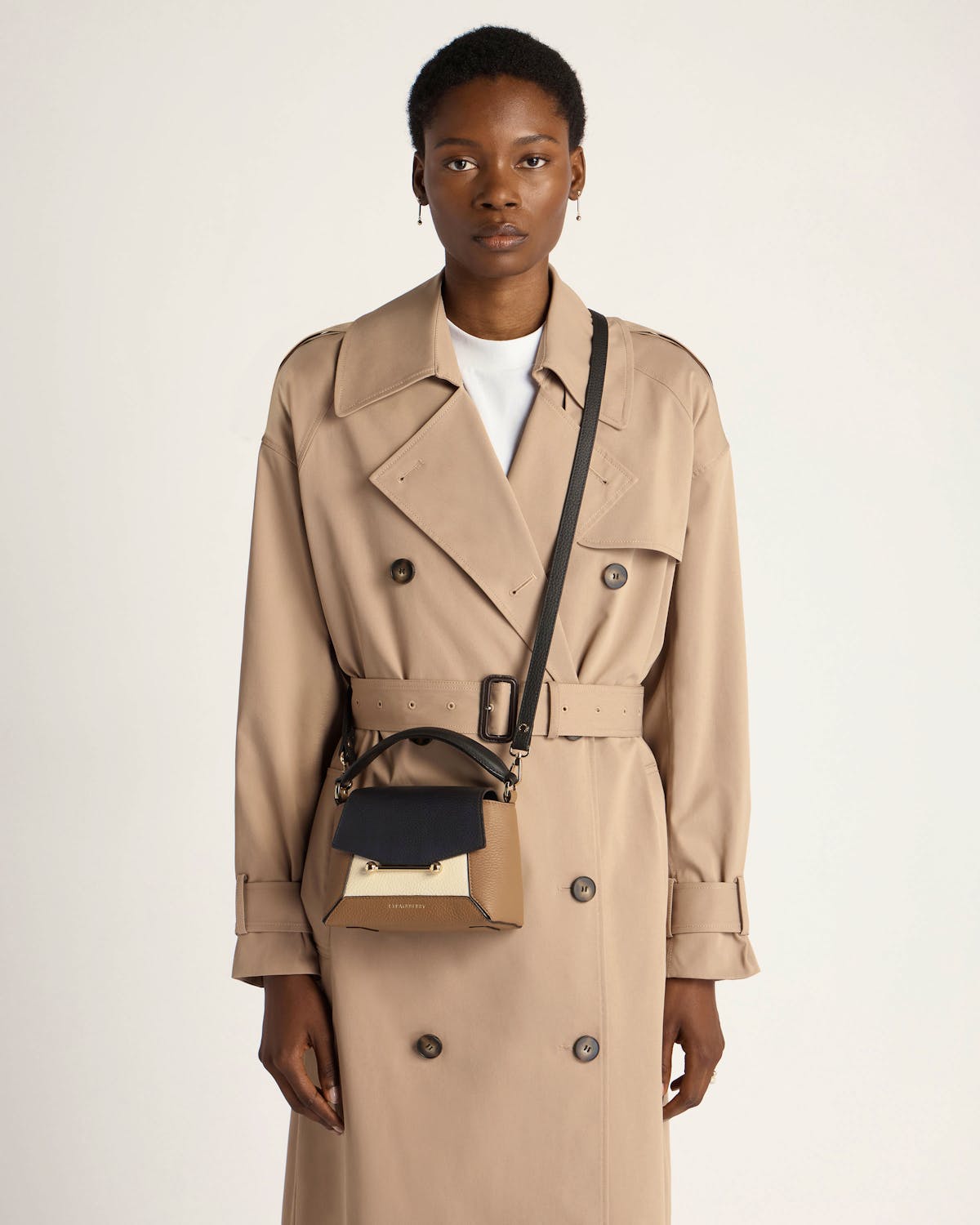 Model wearing beige trench coat with small clay, black and white crossbody bag.