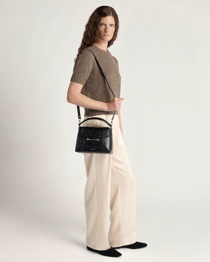 Model wearing brown jumper and cream trousers holding black croc-embossed Mosaic bag.