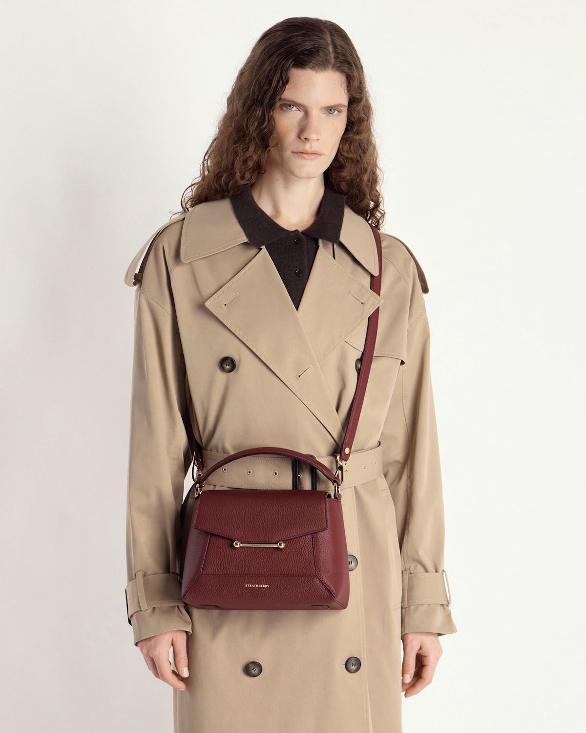 Model wearing a beige trench coat carrying an oxblood mosaic bag by Strathberry crossbody.