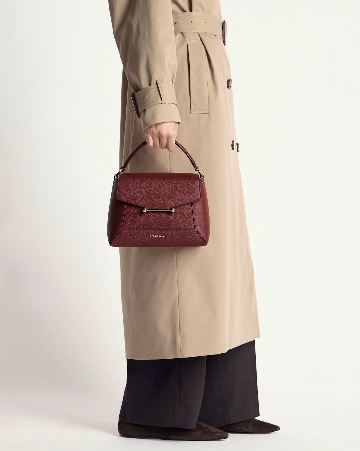 Model carrying oxblood Strathberry Mosaic bag with beige trench coat and black trousers.