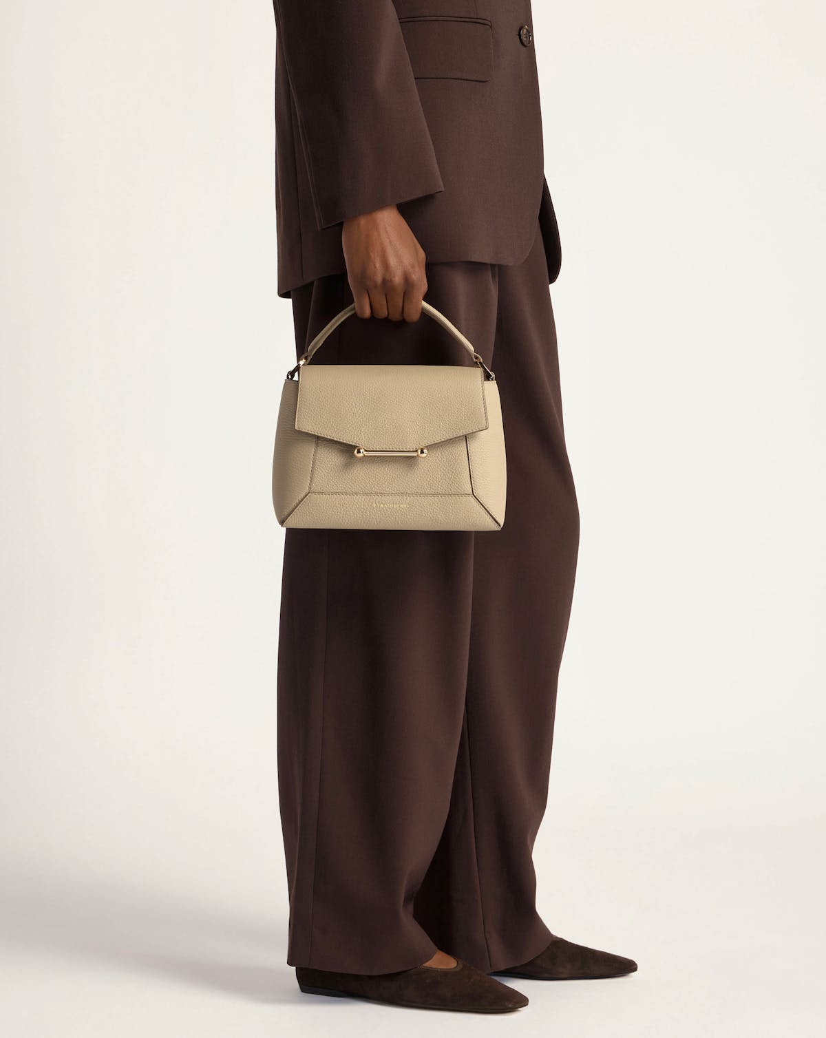 Model holding a sand mosaic bag by the top handle, wearing brown suit and flats.