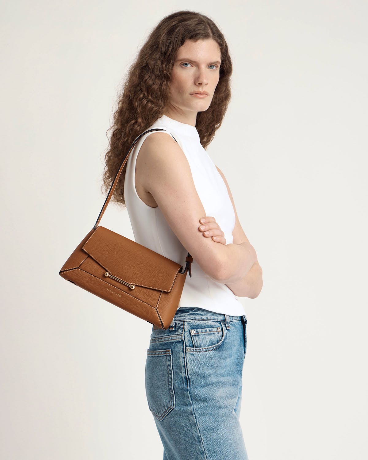 Woman wearing tan leather shoulder bag with flap and gold bar detail, side view.