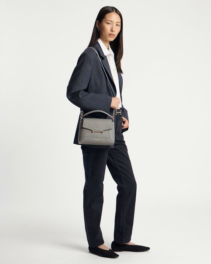 Model wearing dark blazer and trousers carrying a stone grey mosaic bag with strap.