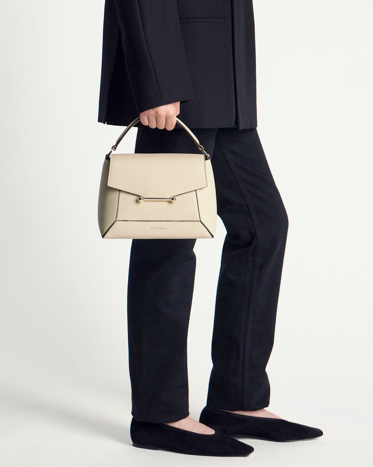 Model holding a vanilla Strathberry Mosaic bag by its handle, wearing black trousers and flats.