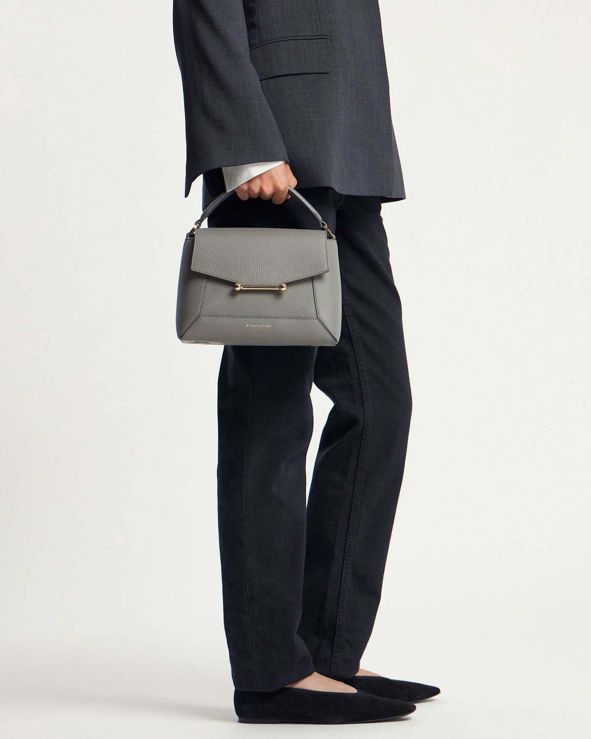 Model holding stone grey mosaic bag by top handle, wearing dark trousers.