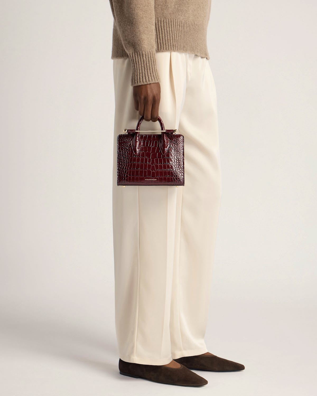Model holding burgundy croc-effect nano tote by Strathberry at her side.