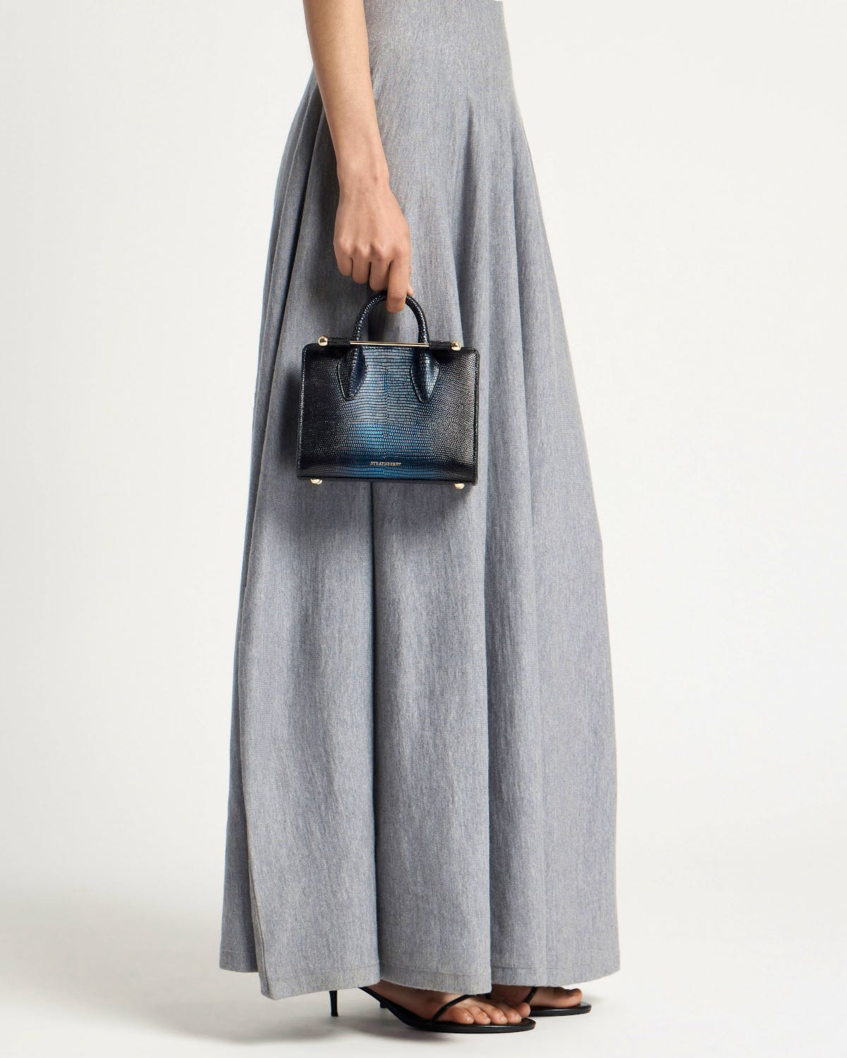 Model holding a small lizard-effect nano tote by its top handles, grey maxi skirt.