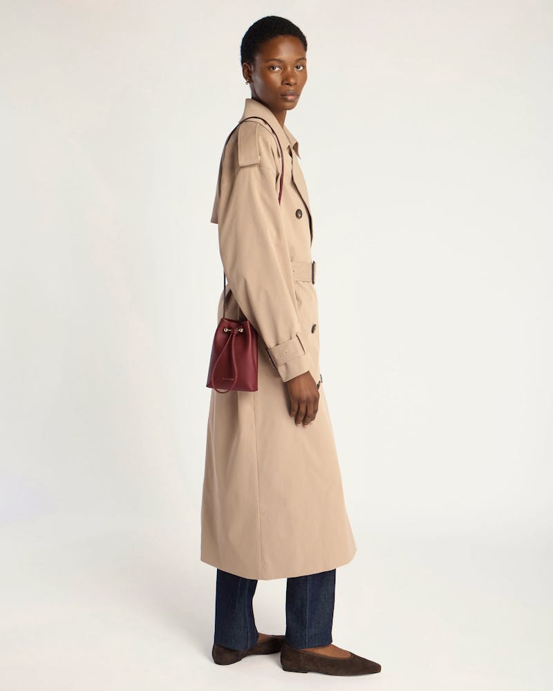Model in beige trench coat wearing oxblood midi pouch on shoulder.