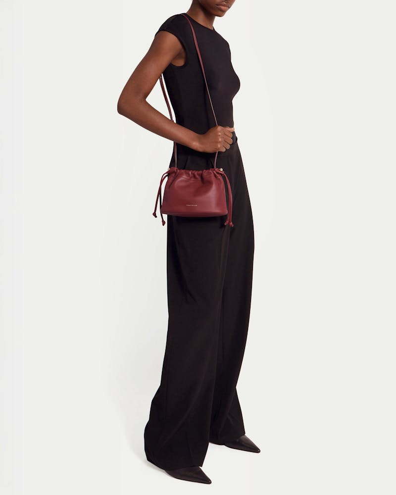 Model wearing black outfit carrying oxblood drawstring shoulder bag worn at hip.