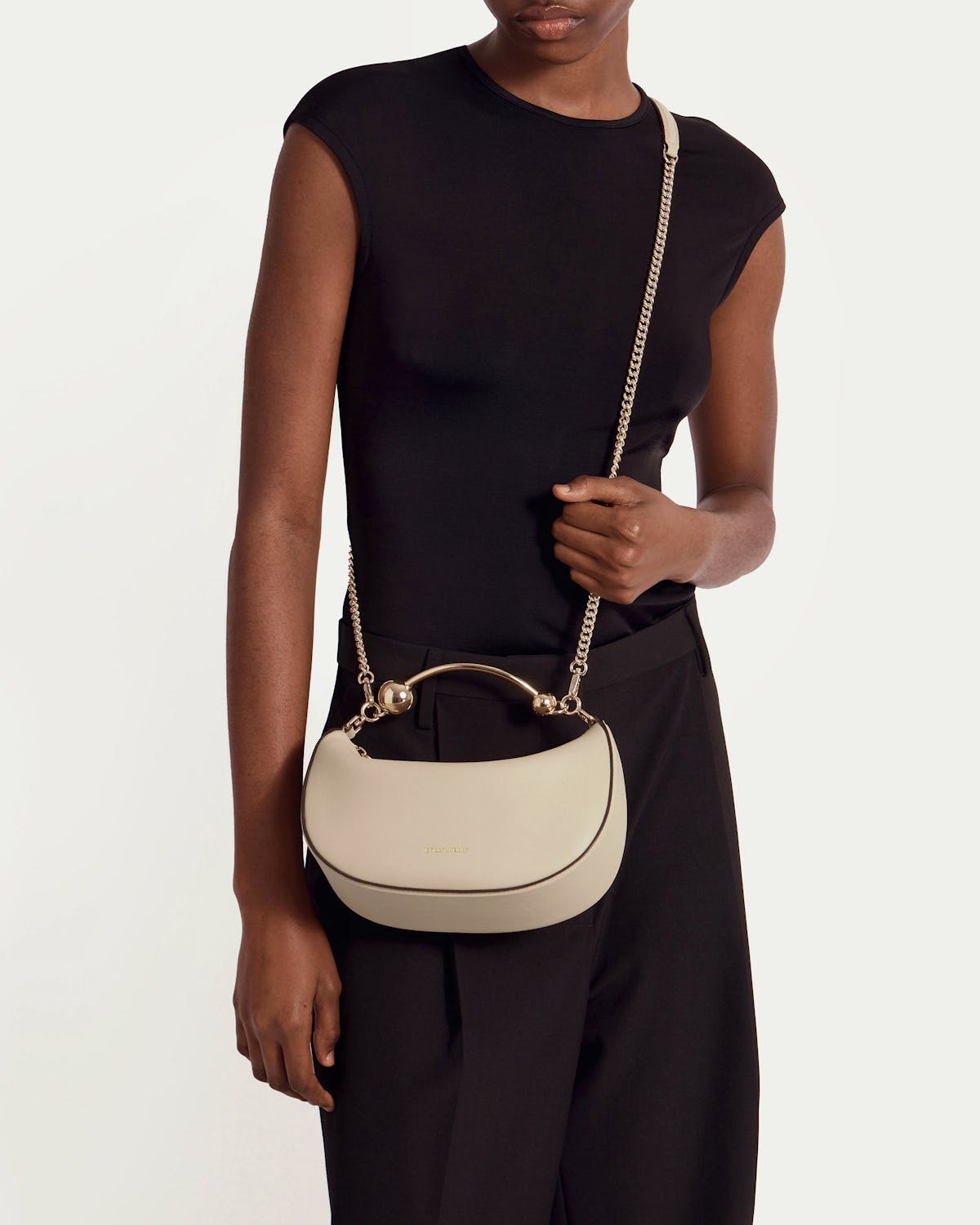 Model wearing oat crescent-shaped mini bag with gold chain strap.