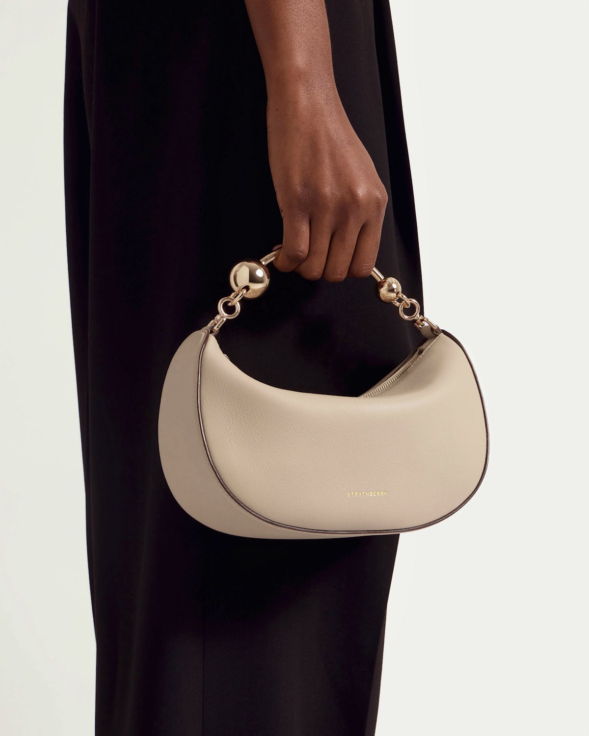 Strathberry Crescent Moon Mini bag in oat leather held by model's hand.