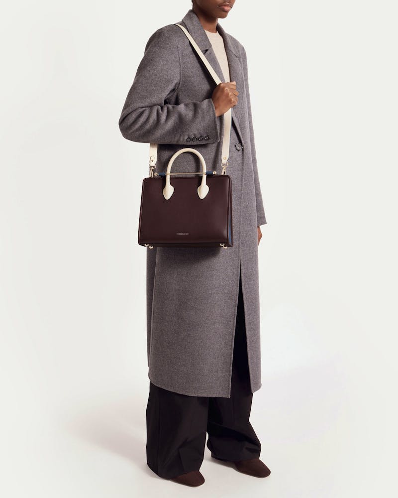 Model in grey coat holding burgundy midi tote with vanilla handles and navy trim.