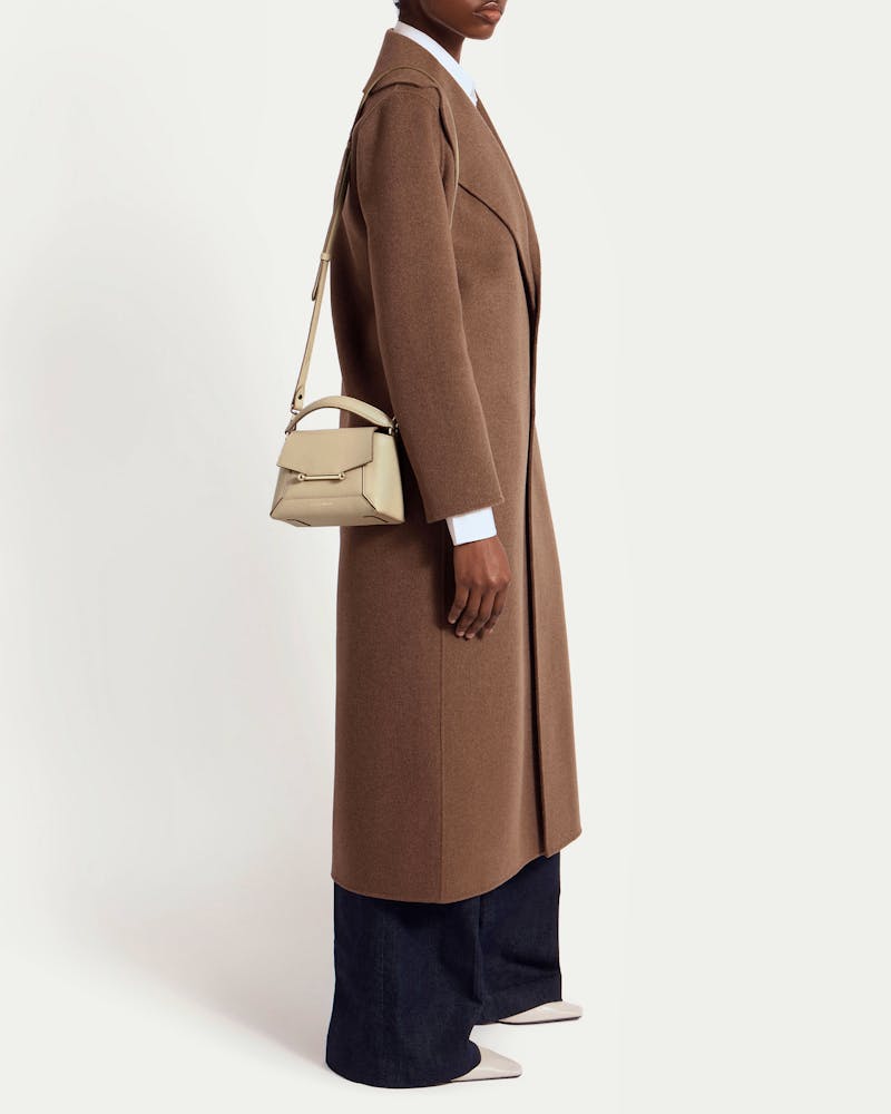 Model wearing a long brown coat with an oat Mosaic Nano shoulder bag.