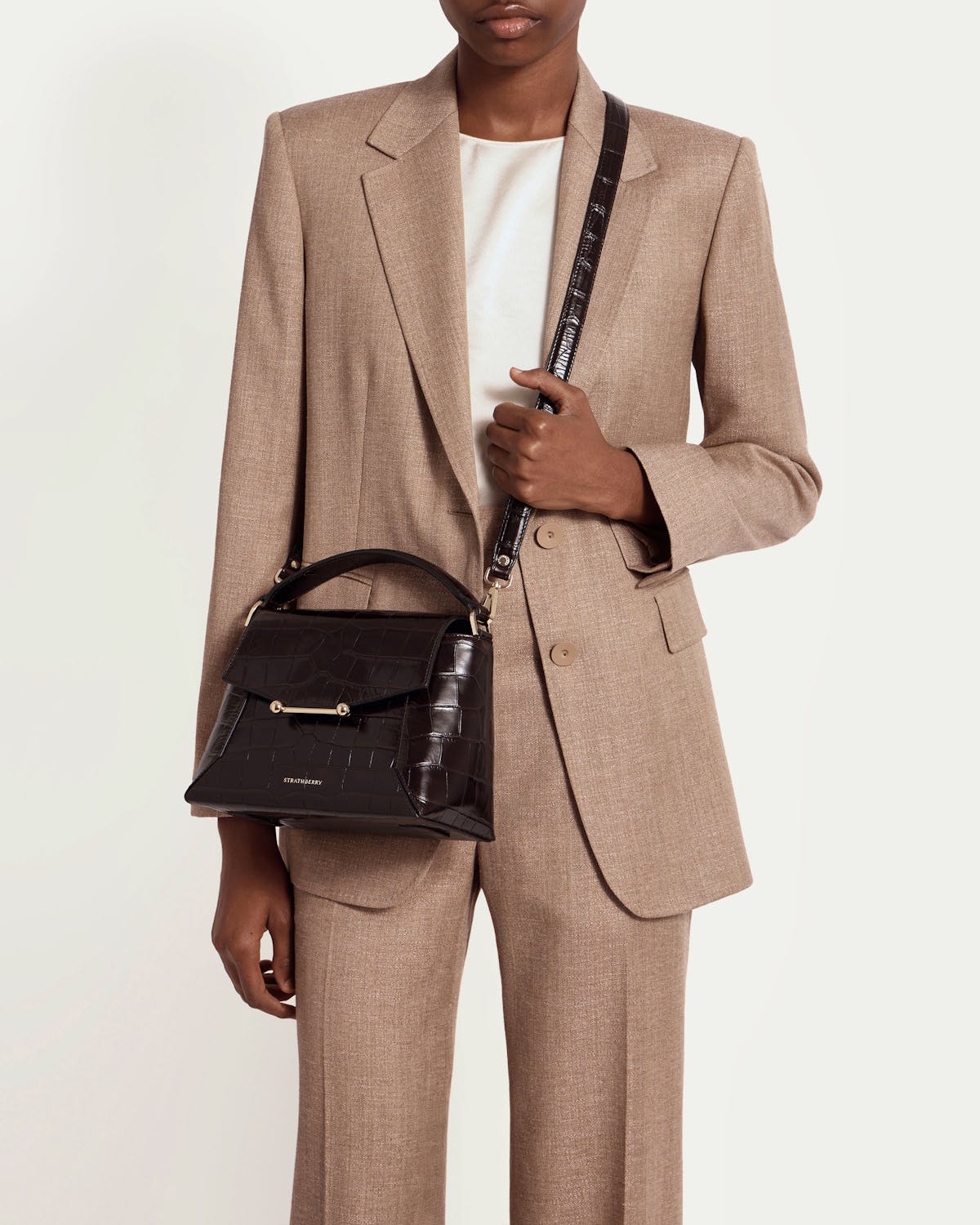 Model in beige suit holding dark croc Mosaic bag with Strathberry logo.